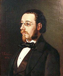 Portrait of a fairly young man in spectacles, with beard, moustache and longish hair, wearing a dress shirt with a large floppy bow tie