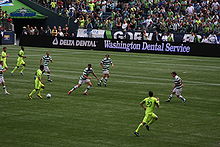 Players advancing on the goal during a soccer match. Banners in front of the crowd read "Gorilla FC".