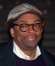 A headshot of a middle aged African American man. Wearing round glasses and a silver cap, the man sports dark stubble.