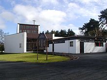 Modernist Church coated with white render with large modern stained glass window and a wooden cross protruding from the roof of the two storey tower element of the building