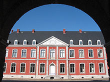 Through the archway of a gate is visible a wide part of the red-brick façade of a two-storey building, with large windows outlines with white stone arches. They grey roof shows skylight windows of a third floor. The entrace is preceded by a small set of steps and is surmounted by an ornament showing two figures supporting a coat of arms.