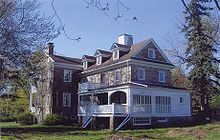 A large stone house with many dormers, windows, and a white porch, surrounded by trees