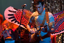 A male vocalist with a banjo performing on stage, wearing large, colourful wings