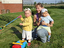 A young mother kneels in a garden with her two children. A baby sits astride her knee facing outwards and looking away from the camera. A toddler stands slightly in front of his mother holding a spade and frowning at the camera.