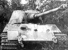 A head-on view of a large tank with a flat faced turret. Its sloped bow armor is scarred with several fist-sized dents, and there is a fist-sized hole in the front of the turret