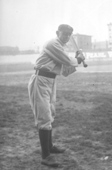 A man wearing a baseball uniform standing truned to his left poised to swing a baseball bat.