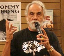  A well-tanned man with white hair and a white beard holds a microphone in front of his face. On his left wrist, he wears a heavy silver-colored watch; with his right hand, he is gesturing. On the wall behind him are two signs: one bears the name "Tommy Chong".