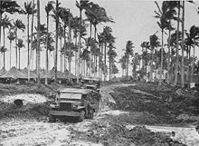 Two World War II-era trucks driving along a muddy road. Tents and palm trees are visible in the background.