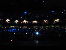 A darkened concert venue. People are in multiple levels of seating, and tiny lights are scattered throughout the audience. On the left, performers stand on a stage lit with blue lighting.