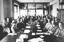 Photo of a caucus meeting showing thirty-five men and a woman, sitting around several long tables with documents laid out before them. As the space is cramped and there is not room for everyone at the table, they are seated in two rows.