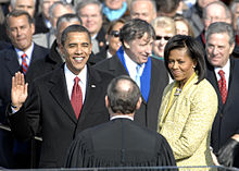 Barack Obama holds his right hand in the air as he and Michelle Obama both smile towards a balding man whose back is to the camera while a large crowd watches.