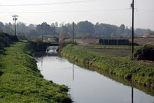 A stream perhaps 20 feet (6 m) wide flows between grassy banks and telephone poles on either side. Waterfowl float on the stream near a low bridge in the middle distance. In the far distance is a line of trees.