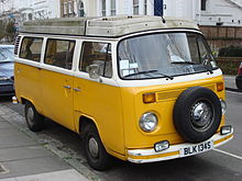 A yellow and white Vokswagen Type 2 vehicle, at center, is parked on a street. The roof of the vehicle is dirty, and on the sides of the picture, several other vehicles and buildings can be seen located on the street.