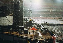 A side shot of a concert stage as a crew disassembles it at night. The stadium it is built in is empty and lit up.