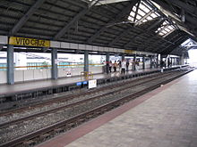 A relatively empty boarding platform with only a handful of people identified by a sign as that at Vito Cruz station. Pebbles line the tracks and sunlight comes in from spaces open to the outside and large open flaps in the dark warehouse-like roof.