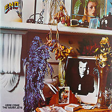 A close up photo of a mantle with a desk below it. Items on the mantle include a color photo of Brian Eno, a kettle and flowers. Items on the desk below are a black-and-white photo of Eno, flowers, playing cards and cigarettes. In the top left corner of the album cover "Eno" is written. At the bottom left corner of the album, "Here Come the Warm Jets" is written.