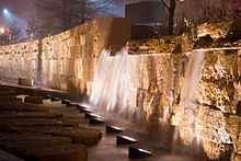 To the right of some stepping stones, a row of lamps illuminates a short artificial waterfall streaming down a honey-colored wall made of limestone bricks.