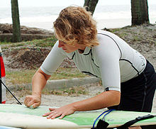 Photo of man bent over surfboard rubbing bar of solid wax against the board with palm trees and ocean in background