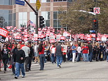 A large group of people in front of a building, picketing with signs reading "Writers Guild of America on Strike".