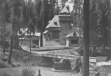 A monochrome photograph of a 75-foot-tall building with steeply pitched roof elements, seen amid tall pine trees from across a river. A rustic one-lane bridge over the river is in the foreground.