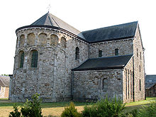 A small stone church stands in a field. The apse shows two small stained-glass windows, with empty arched niches above. The left transept is also visible, with a half-height chapel adjacent.