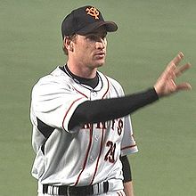 A white man in a black baseball cap and a black long-sleeved shirt with a white baseball jersey over it extends his right hand.