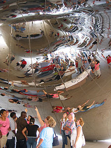 The reflections of visitors are warped in the mirror-like surface of an object above them; the surface of the object is covered in weld lines.
