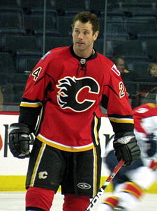A man with short, brown hair looks to his right.  He is wearing a red uniform with black pants and a black stylized C logo on his chest.