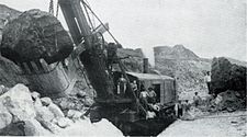 A black and white photograph showing part of another, similar steam shovel in an area of disturbed earth with a large rock in the bucket