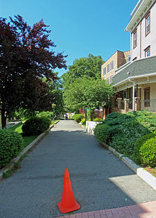 An asphalt path with an orange traffic cone at its beginning, in front of the camera. There are buildings and trees on both sides.