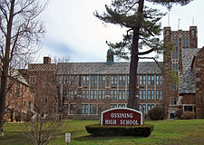 A complex two-story brick building with a tower, cupola and multicolored roof. In front are a pine tree and some bare ones, along with a white-on-maroon sign reading "Ossining High School".