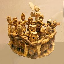A ring of twelve dancing figures, arms interlocked around each others' shoulders. They surround one musician in the centre of the ring, and a second musician stands behind them.
