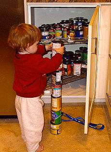 Young red-haired boy facing away from camera, stacking a seventh can atop a column of six food cans on the kitchen floor. An open pantry contains many more cans.