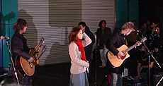 A male guitarist on the left, a female singer in the center, and another male guitarist on the right performing on the street with several people behind them