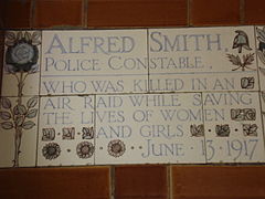 A tablet formed of five tiles of varying sizes, elaborately decorated with flowers and a stylised English policeman's helmet and bordered by yellow and blue flowers in an art nouveau style. The tablet reads "Alfred Smith, Police Constable, who was killed in an air raid while saving the lives of women and girls June 13, 1917".
