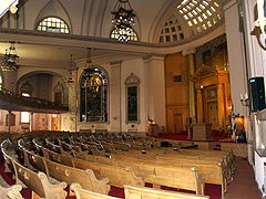 The front of a wide, two to three-story room is visible. Five rows of wooden pews narrow to three as they approach the front wall, which has a large protruding wooden Torah ark at its center. To the left of the ark is a large, arched stained-glass window. The edge of a second-floor balcony which projects partway into the sanctuary is visible on the left side of the picture. The ceiling is arched, with multi-paned stained-glass windows in it, and elaborate chandeliers with Star-of-David shapes hanging from it.