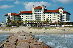 A stately white hotel building with a red-tile roof is seen from the end of a jetty extending from the beach.