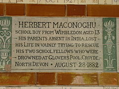 A tablet formed of six standard sized tiles, bordered by green flowers in the style of the Arts and Crafts movement. The tablet reads "Herbert Maconoghu schoolboy from Wimbledon aged 13. His parents absent in India, los his life in vainly trying to rescue his two school fellows who were drowned at Glovers Pool, Croyde, North Devon August 28, 1882".