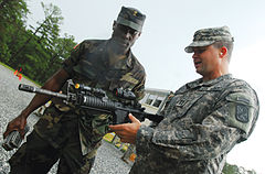 A colour photograph of two soldiers, one holding an assault rifle, talking at a training ground. The soldier on the left is in a camouflage field uniform, while the soldier on the right holding the rifle is in a grey camouflage uniform
