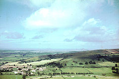 The houses and church of a small village can be seen in the bottom left of the picture. It is surrounded by a patchwork of fields with some trees on a hillside. Large hills in the distance.