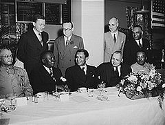 A number of business-suited and uniformed men sit facing the camera along a table