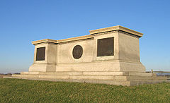 A large monument of off-white stone.  Its shape resembles a long, flat pedestal.  It sits atop a grassy knoll with a bright blue sky overhead.