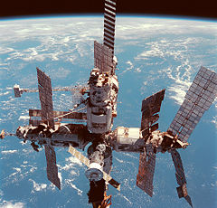 A view of Mir backdropped by the limb of the Earth. In view are four cylindrical modules covered in white insulation arranged in a cross shape about a small, central sphere. Another module projects backward from this sphere, and a small module is attached to the far end of that. Each module is sprouting various solar arrays, cranes and other spindly equipment, with Soyuz and Progress spacecraft docked to the forward and aft ports of the complex.