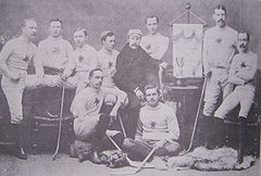 A group of men wearing their team uniforms, some standing and some sitting, each with a hockey stick, in a studio. On an easel is a Ottawa championship banner. On a table is a trophy.