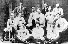 Ten men, wearing white team uniforms, in two rows across the picture, each holding a hockey stick. Behind them are two men, team executives, standing in their suits, one wearing a bowler hat.