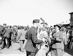 Man in dark military uniform in the arms of a female civilian, among a crowd of other civilian and military people