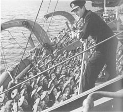 A man in a naval uniform addresses soldiers from a platform on a ship