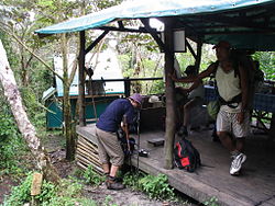 Several people prepare for a hike under a covered kitchen area, with a bungalow visible in the background.