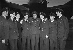 Portrait of a group of eight men in formal military uniform. All are wearing caps, and have their arms around each other. A large aircraft is in the background.