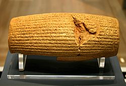 Front view of a barrel-shaped clay cylinder resting on a stand. The cylinder is covered with lines of cuneiform text